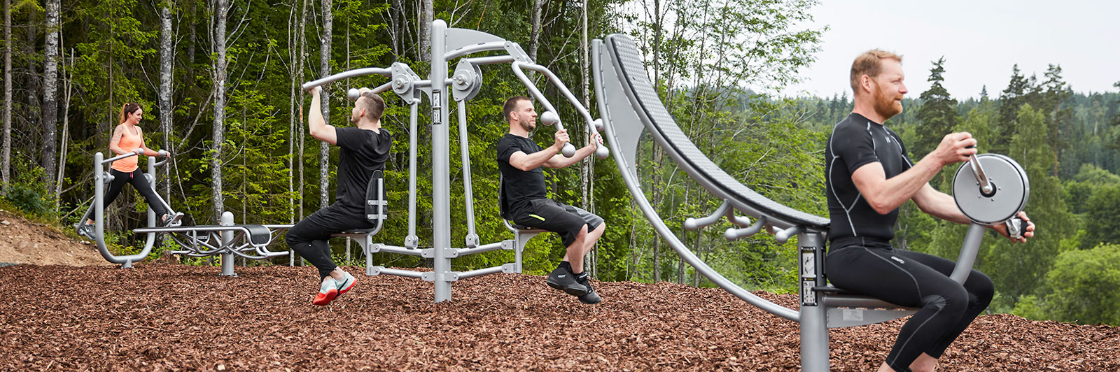 A group of adults exercise on various outdoor fitness gym equipment among nature.